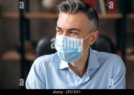 Primo piano di un uomo caucasico di successo, manager o lavoratore d'ufficio, leader di affari che indossa una maschera medica, in una camicia elegante, guarda via. Prevenzione, precauzioni e protezione Foto Stock