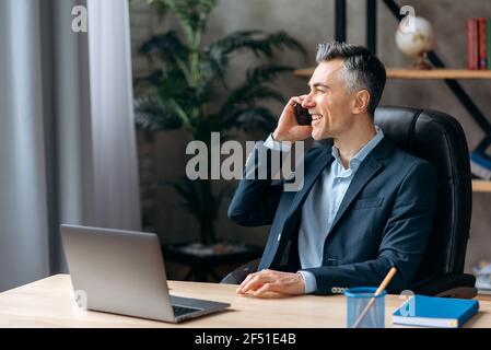 Riuscito agente immobiliare adulto caucasico sicuro di sé, seduto in ufficio moderno, in abito elegante, parlando su un cellulare con un cliente, guardando via, sorridente amichevole Foto Stock