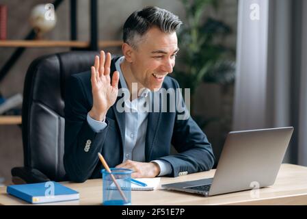 Influente successo attraente caucasico maturo ceo, business leader o manager, in tuta formale, comunica tramite videochiamata, conduce online briefing con i dipendenti, Waves mano, sorrisi amichevole Foto Stock