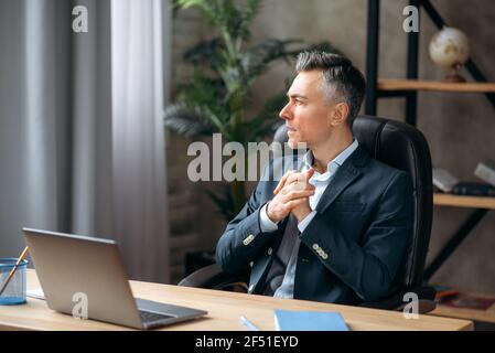 Bell'auto-sicuro successo medio-vecchio leader o manager caucasico di affari, seduto in un ufficio in un elegante tuta formale, pensando a un progetto, guardando via, prendendo appunti, sorridendo Foto Stock