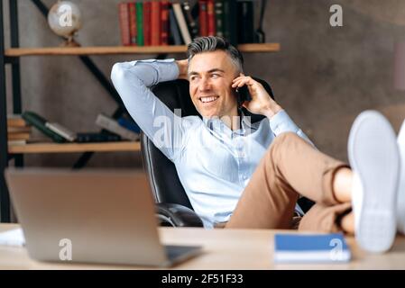 Leader, freelance o manager caucasico, con i capelli grigi, influente e fiducioso, getta i piedi sul tavolo, parla al telefono, chiama amici o colleghi, guardando via, sorrisi felici Foto Stock