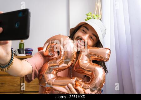 Attraente giovane latino-asiatico prendendo un selfie da solo, tenendo un pallone d'aria metallico, festeggiando il suo 25° compleanno. Palloncino metallico con numero Foto Stock