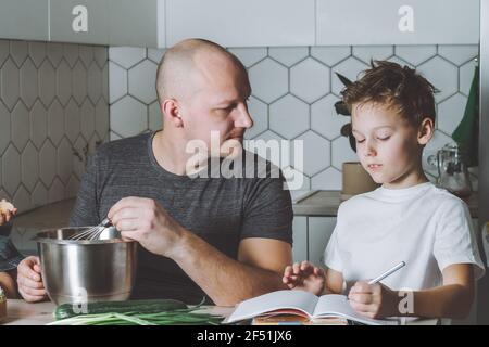 Padre fruste omelette e aiuta suo figlio a fare i suoi compiti in cucina. Uomo che fa le faccende domestiche. Foto Stock