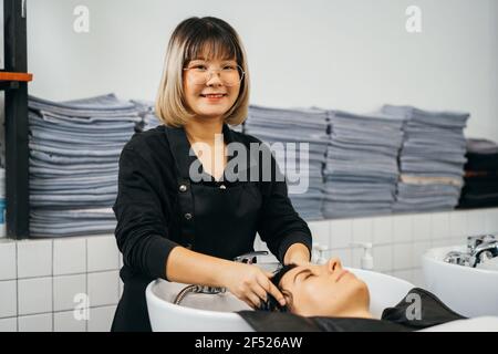 Ritratto di giovane estetista asiatica guardando la macchina fotografica, in piedi e massaggiando i capelli e la testa di giovane cliente femminile con la testa in lavabo sorridente e guardando la macchina fotografica Foto Stock