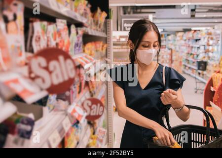 Bella giovane donna asiatica che indossa covid-19 maschera protettiva shopping e mettere il pacchetto di cibo in paniere in ipermercato Foto Stock