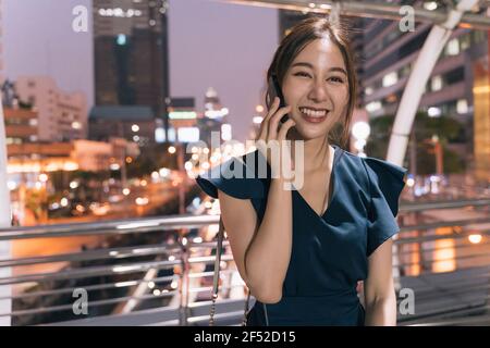 Ritratto di sorridente giovane asiatica riuscita donna in abbigliamento casual parlare sullo smartphone mentre ci si trova sul ponte pedonale con vista sulla città in background Foto Stock