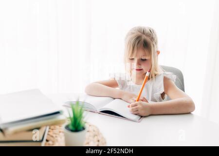 Una bambina si siede al tavolo e scrive in un taccuino bianco. Concetto di istruzione. Scuola domestica. Compiti. Foto di alta qualità Foto Stock