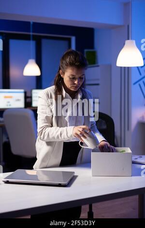 Depresso dipendente licenziato mettendo il suo roba in scatola bianca dal tavolo in ufficio come essere licenziato. Disoccupato che imballano le cose a tarda notte. Donna che lascia l'ufficio del posto di lavoro a mezzanotte Foto Stock