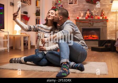 Felice coppia sorridente mentre prende un selfie il giorno di natale. Gatto dormendo sullo sfondo. Foto Stock