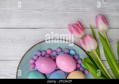 Uova pasquali di colore pastello e caramelle di cioccolato di colore pastello in una piastra di colore blu e tulipani bianchi rosa, su sfondo di legno bianco. Foto Stock