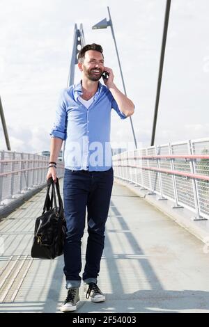 Ritratto a lunghezza intera di felice uomo maturo che cammina sul ponte e utilizzando il telefono cellulare Foto Stock