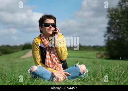 Donna che indossa occhiali da sole, cappotto giallo e jeans seduti sul prato in una giornata soleggiata e calda nel parco Foto Stock