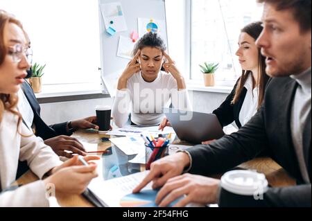 Uomini d'affari multirazziali in riunione di briefing avendo un brainstorming teso. I colleghi che hanno lavorato troppo non possono trovare idea per il progetto o venire d'accordo, sentendosi esausti e frustrati Foto Stock