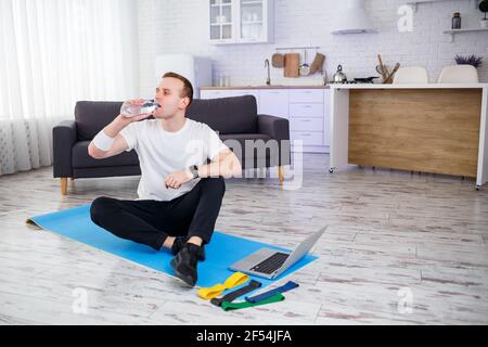 Felice giovane uomo beve acqua dopo l'allenamento online. Fare sport a casa, uno stile di vita sano Foto Stock