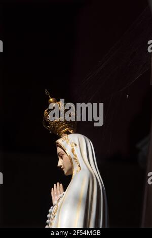Pregando la statua di Madonna Fatima con un ciottolo sulla sua corona in una vuota chiesa scura. Background religioso. Concetti di misericordia, miracolo, speranza, preghiera. Foto Stock