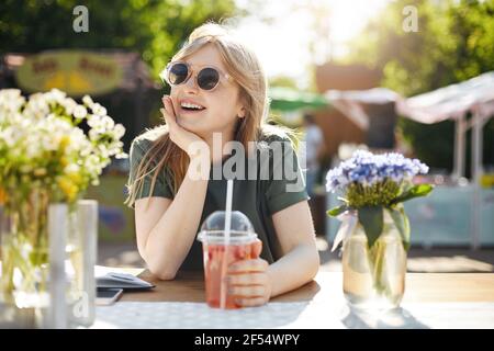 Ritratto di giovane adorabile donna che beve limonata in parco surrondato da fioriture, indossando occhiali sognando della sua carriera futura come blogger beaty. Foto Stock