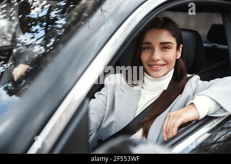 Donna felice ed elegante seduta in auto con cintura di sicurezza allacciata, guida al lavoro. Il driver femmina sorride alla telecamera, guardando fuori dalla finestra Foto Stock