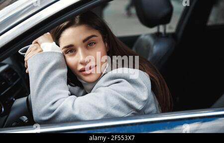 Bella ed elegante donna driver riposante nella sua auto, appoggiata sulla ruota dell'auto e guardando fuori finestra, in attesa nel parcheggio Foto Stock