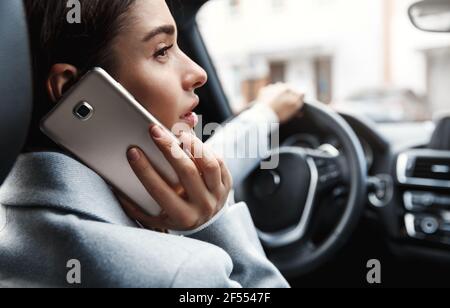 Primo piano di una giovane donna d'affari che guida in ufficio e chiama qualcuno al telefono. Donna seduta sul conducente seduto e parlando sullo smartphone Foto Stock