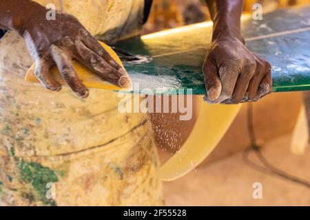 Uno Shaper da surf di un artigiano africano che lavora in un'officina di riparazione Con carta vetrata Foto Stock