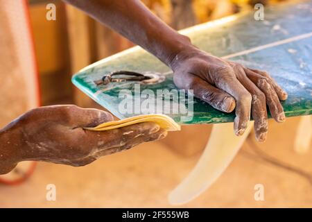 Uno Shaper da surf di un artigiano africano che lavora in un'officina di riparazione Con carta vetrata Foto Stock