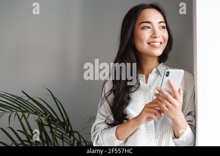 Ritratto di bella donna asiatica moderna guardando fuori finestra e sorridente sognante, tenendo smartphone. Ragazza in attesa di una chiamata telefonica, telecomando funzionante Foto Stock