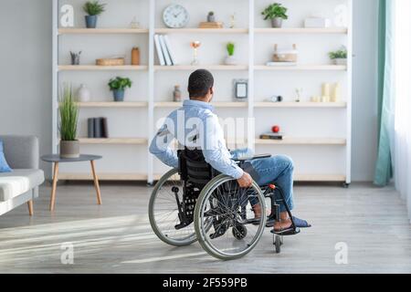 Depressione negli adulti con disabilità. Infelice giovane uomo nero in sedia a rotelle sentendosi solo a casa, a tutta lunghezza Foto Stock