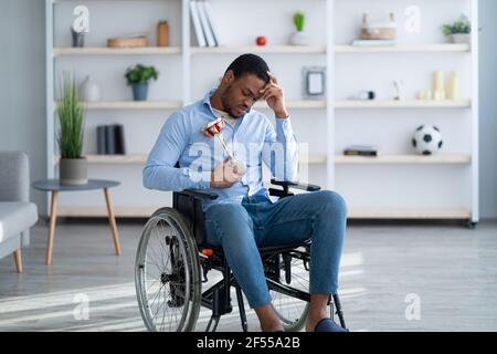 Ritratto di triste sportivo nero con trofeo seduto in sedia a rotelle, sentendosi depresso per la sua ferita a casa Foto Stock