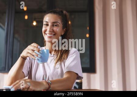 Bella donna sorridente che beve il tè sulla sua terrazza di casa, tenendo la tazza e guardando felice alla macchina fotografica, seduto al tavolo da caffè in t-shirt Foto Stock