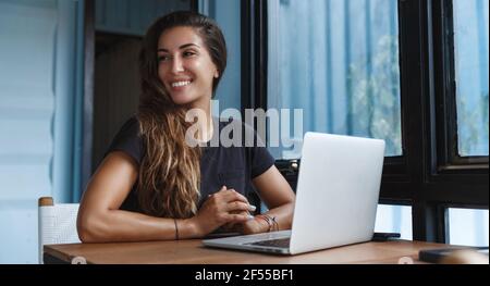 Donna freelance che lavora da casa, seduto a tavola con il computer portatile e guardando da parte con sorriso felice. Ragazza indiana che ha riunione in linea sul calcolatore Foto Stock