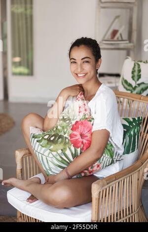 Foto verticale di felice donna tenera seduta nel patio accogliente dell'hotel, balcone, gambe trasversali abbraccio carino cuscino, riposo in poltrona di rattan, per prepararsi Foto Stock