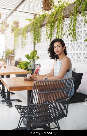 Foto verticale sognante attraente donna in attesa di qualcuno tavolo da caffè seduto l'entrata del ristorante si trova in una veranda e offre un check-out curioso Foto Stock