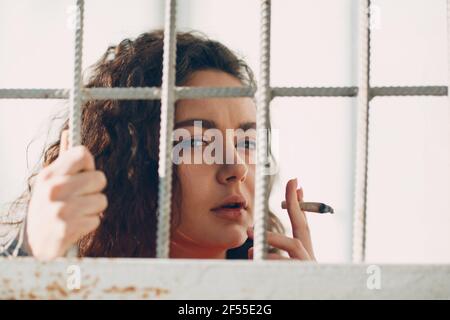 Giovane brunetta donna riccia in tuta arancione dietro bar di prigione fuma marijuana giunto. Donna in coloratissima tuta ritratto Foto Stock