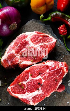 Bistecche di carne crude su sfondo nero con verdure, cipolle azzurre fresche, peperoni gialli, peperoncino. Immagine di sfondo. Vista laterale, vista dall'alto Foto Stock