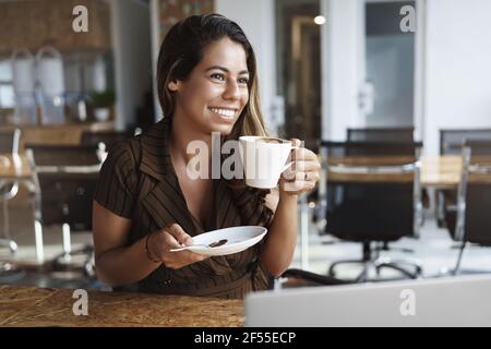 La donna gode di un gustoso caffè nel caffè durante il pranzo. Attraente imprenditore femminile che lavora a distanza in co-working, tenere tazza e piatto, sorridente felice come Foto Stock