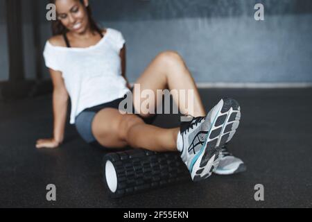 Donna seduta sul pavimento della palestra in activewear, t-shirt bianca, faccia offuscata come fuoco sulle gambe che fanno l'esercitazione con l'apparecchiatura funzionale di addestramento, rullo della schiuma Foto Stock