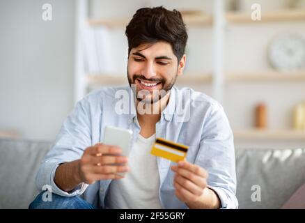 Acquisti online veloci. Uomo arabo felice usando il telefono e la carta di credito per ordinare i prodotti in Internet Foto Stock