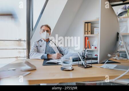 Medico maturo che indossa una maschera protettiva seduta alla scrivania studio medico Foto Stock