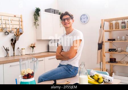 Sorridente giovane seduto con le braccia incrociate sul tavolo dentro cucina Foto Stock