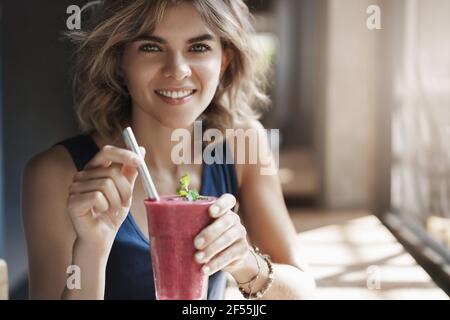 Primo piano sano attraente elegante bionda europea riccia-capelli donna sorridente ampiamente siediti al tavolo vicino alla finestra tenere vetro paglia bevanda frullato Foto Stock