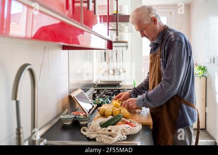 Uomo anziano che trita patate crude su tagliere in cucina Foto Stock