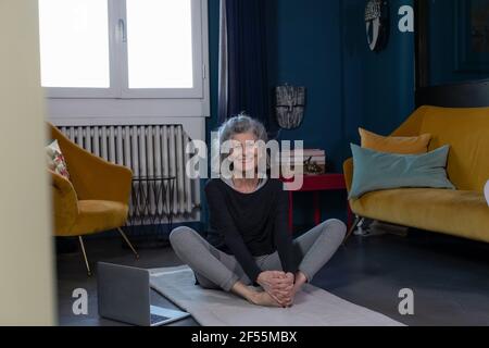 Donna anziana sorridente che pratica lo Yoga sul tappetino da ginnastica a casa Foto Stock