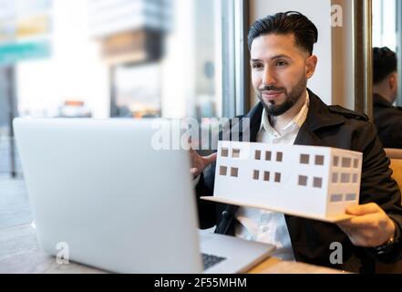 Giovane architetto maschile che mostra il modello architettonico durante la videochiamata computer portatile mentre si è seduti al bar Foto Stock