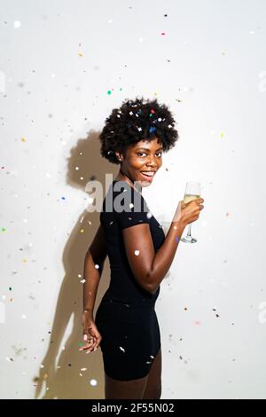 I confetti cadono su una donna allegra che tiene flauto di champagne mentre si è in piedi su sfondo bianco Foto Stock