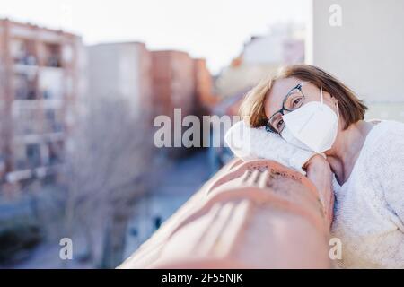 Donna con maschera protettiva appoggiata su ringhiera in balcone A casa durante il COVID-19 Foto Stock