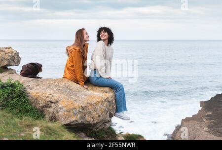 Le amiche sorridono guardandosi mentre si siedono scogliera sul mare Foto Stock