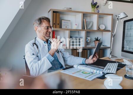 Medico maturo che parla di videochiamata su tablet digitale mentre seduto all'ufficio del medico Foto Stock