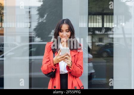 Donna sorridente in trench rosso utilizzando smartphone mentre in piedi contro parete di vetro Foto Stock