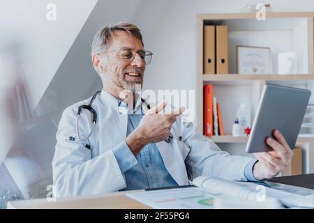 Un medico sorridente che parla di videochiamata su tablet digitale mentre seduto in clinica Foto Stock