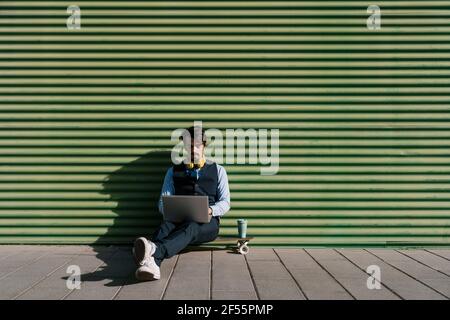 Uomo d'affari che usa il computer portatile mentre si siede sullo skateboard contro la parete verde Foto Stock